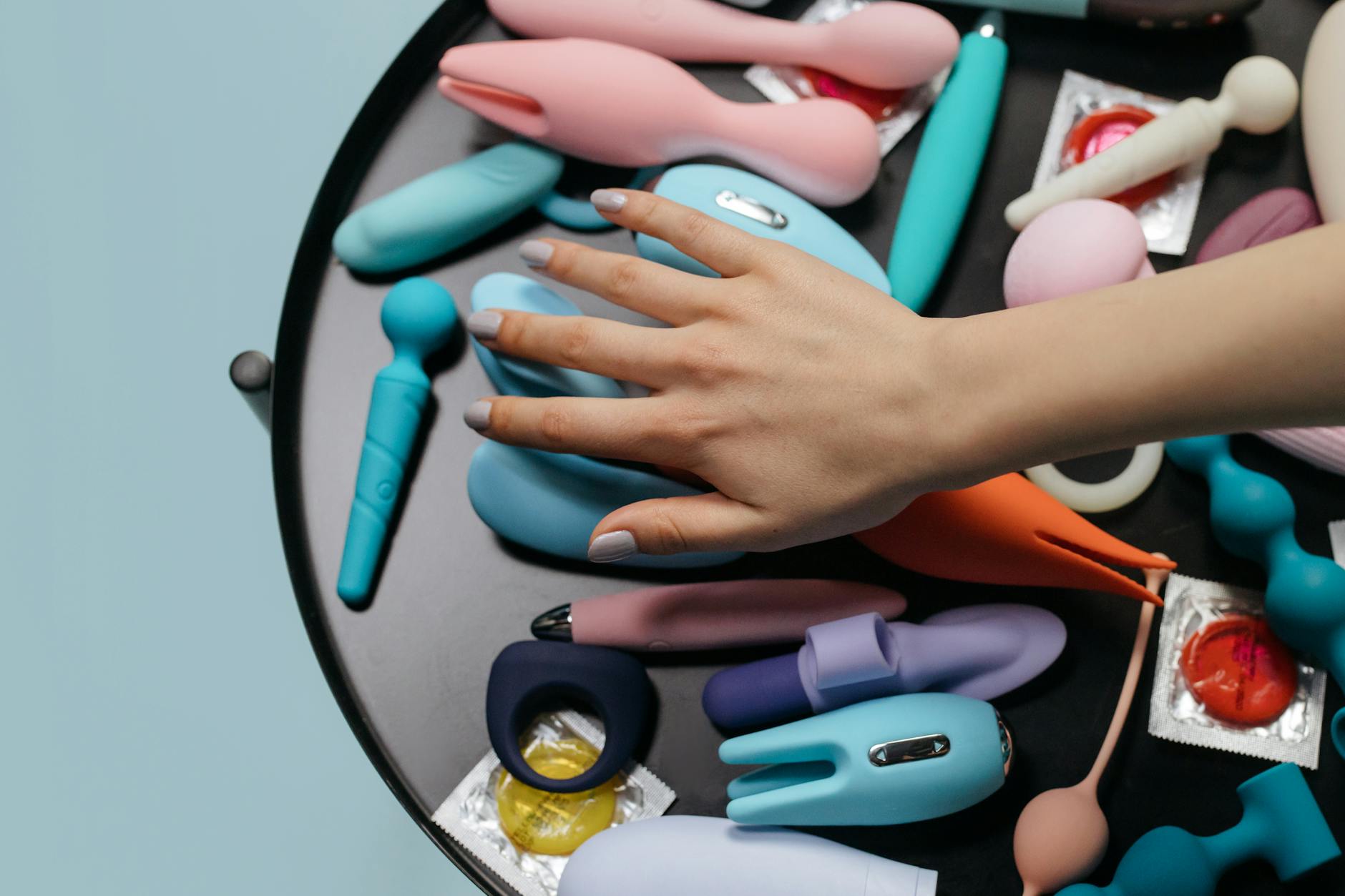 A hand reaching over a variety of colorful sex toys arranged on a table, including lemon-shaped clitoral vibrators and other designs