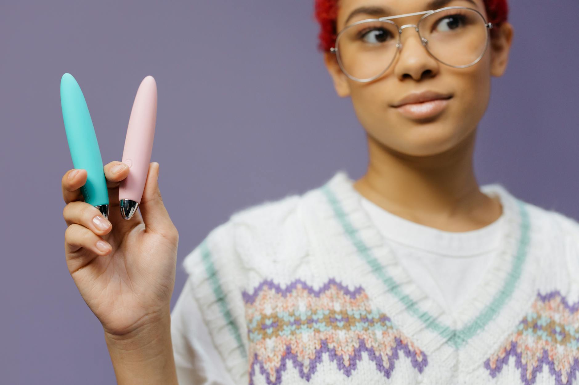 Two hands holding pink and blue silicone vibrators against a pastel background.